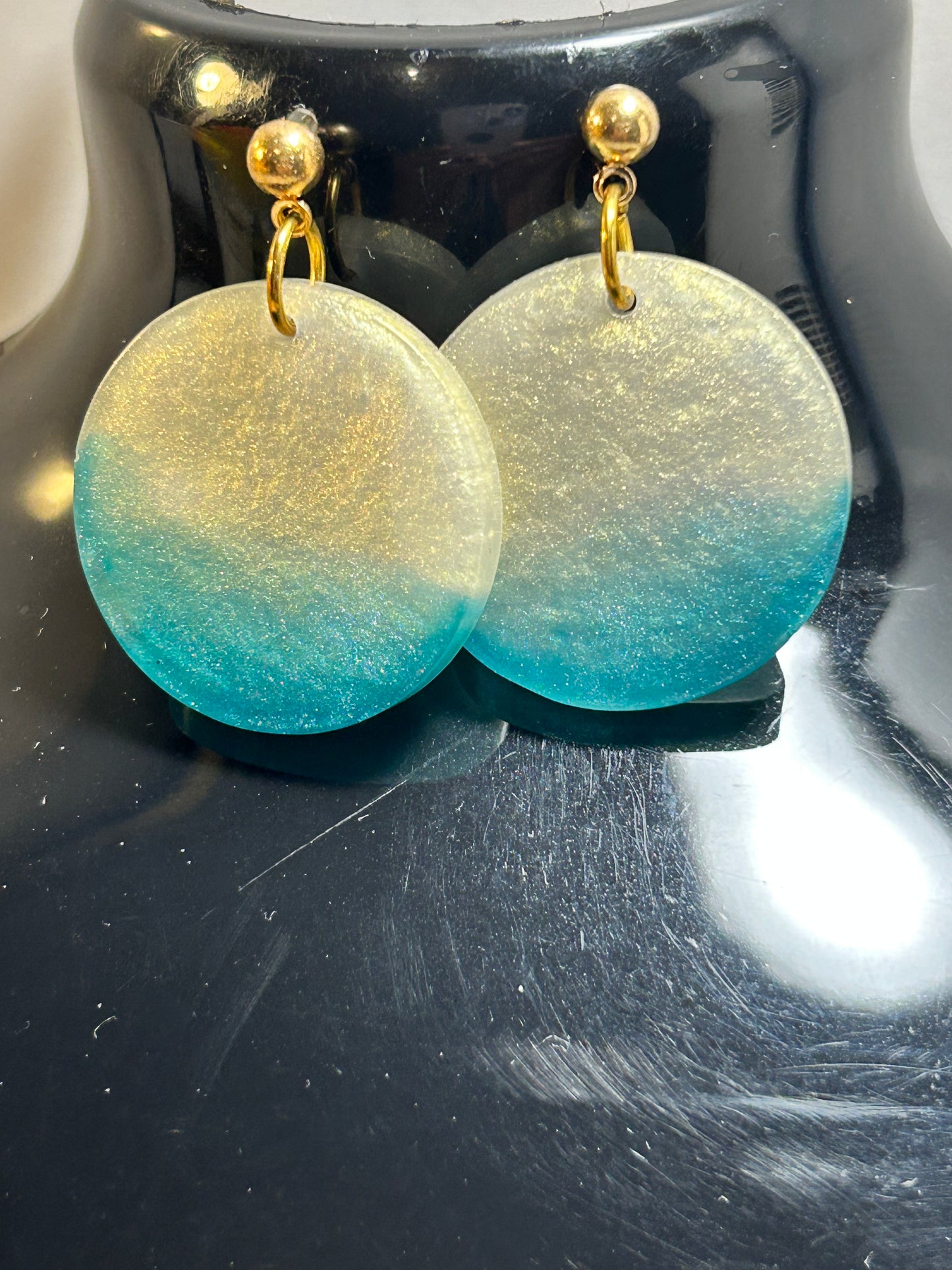 Earrings with gold posts and gradient green to blue disc-shaped pendants on a dark background.